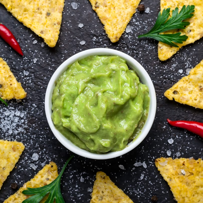 A white bowl of guacamole surrounded by tortilla chips, red chilies, and parsley on a dark surface.