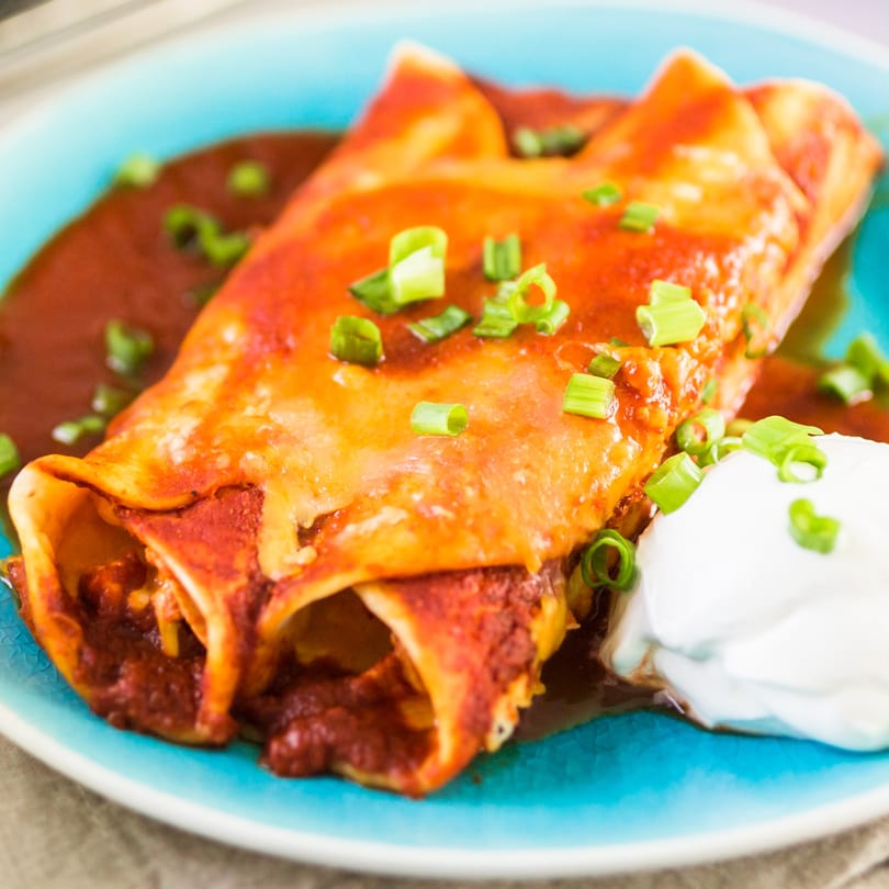 Two Dos Fuentes enchiladas covered in red sauce and green onions, with a dollop of sour cream.