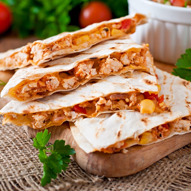 Stacked chicken quesadillas with melted cheese, corn, and red peppers on a wooden board.
