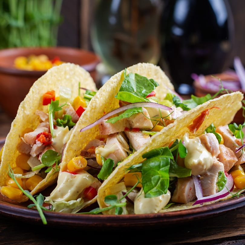 Three Dos Fuentes chicken tacos with corn, red onion, and creamy sauce on a brown plate.