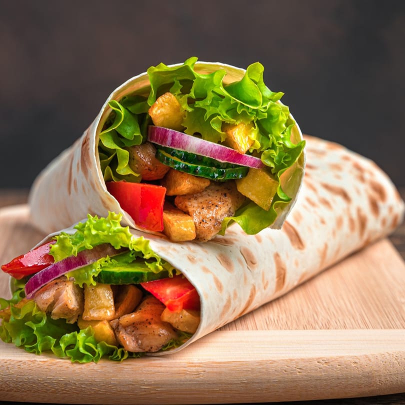 Two Dos Fuentes chicken wraps filled with lettuce, red onion, cucumber, and tomatoes on a wooden board.