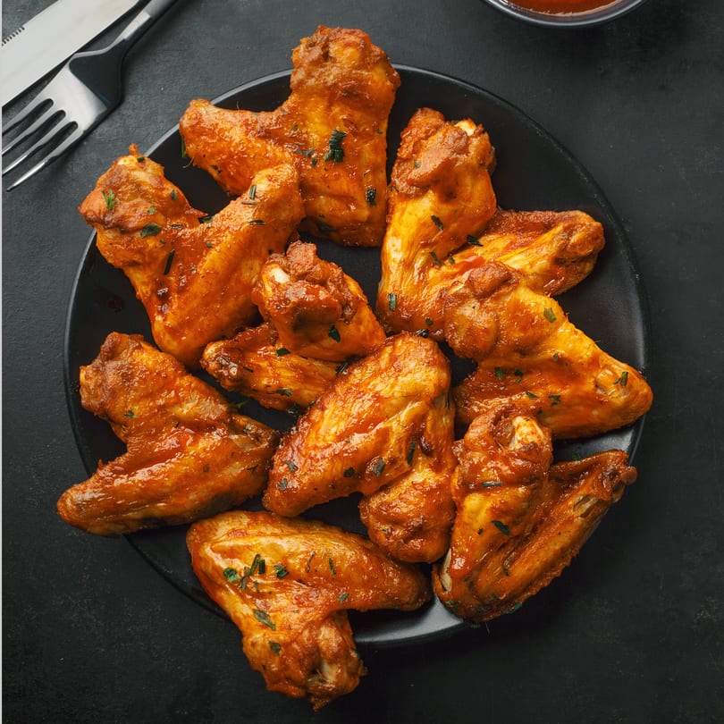 A black plate filled with spicy, glazed chicken wings, garnished with herbs, on a dark surface.